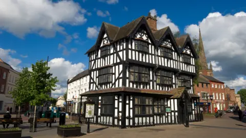 A three-storey tudor building, surrounded by shops. The sky is blue with clouds and there is a church steeple in the right-hand corner of the image.  