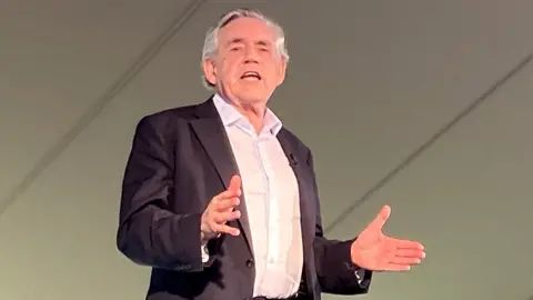 BBC Gordon Brown speaks on stage with his palms outstretched.