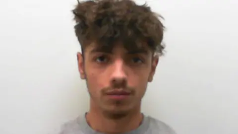 A police mugshot of Kenan Joseph, a man in his twenties with unkempt brown hair, a moustache and a grey sweatshirt.