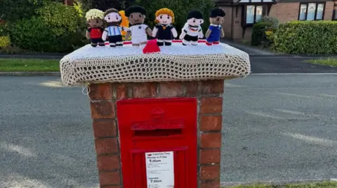 Robyn Cook Knitted footballers in Spanish, Argentinian, English, Scottish German and French team colours line up on top of a crocheted white cover on a red and brick post box. Houses surrounded by bushes can be seen behind.