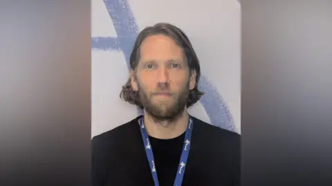 MIND Man with fair hair and a beard wearing a lanyard around his neck. He has a dark jumper on. Behind him is a blue and white background.