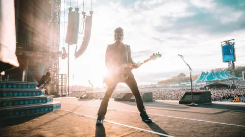 Guns N' Roses A bass player on stage, looking at the camera. Behind him is a large crowd. He is standing with his legs apart, and the sun behind him is dazzling.