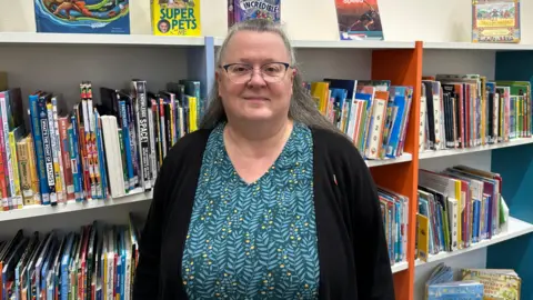 Jayne Cardew is smiling at the camera and has full shelves full of books behind her. She has long hair pinned back from her face and is wearing glasses.