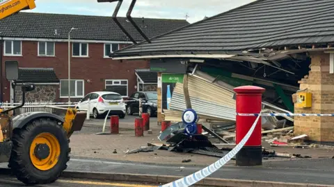 A police cordon around a small village shop which has been rammed into. There is a large hole in the entrance where an ATM would be and the shutters have been torn down.