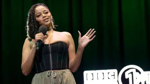 1Xtra Kyrah Gray performing on stage for the BBC 1Xtra Comedy Gala at the Hackney Empire on 1 April. Kyrah is a black woman in her 20s with brown eyes and dark hair worn loose with a platinum blonde streak. She wears a black corset-style top paired with camo-green trousers. She holds a microphone with her right hand while her left is extended out to her side. The staging behind her is dark with BBC Radio 1Xtra branding in lights.