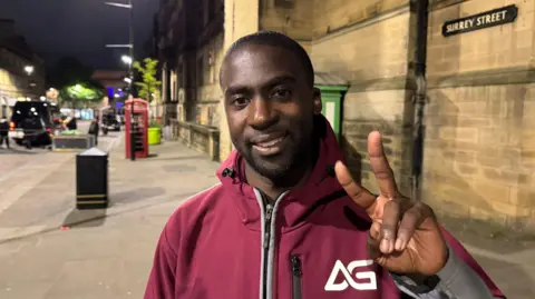 A man stands in a street, wearing a red coat with the letters "AG" on his chest. He holds up his hand in a peace sign.