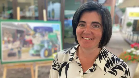 Abi Reader, president of NFU Cymru, smiles at the camera. She has dark, chin-length hair and wears a white blouse with black leaves on it.