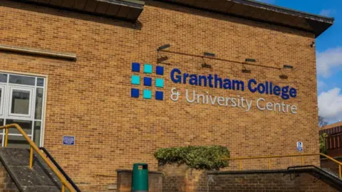 Grantham College The side of a brick building with large words 'Grantham College & University Centre' written in blue and silver. You can see steps leading up to white doors on the left and a green bin in the middle.