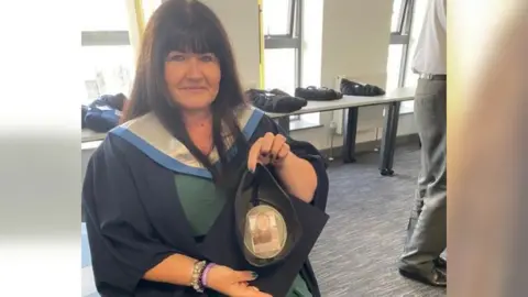 Tanya Russell A women with dark hair holding up a graduation cap with a photo of a young girl inside