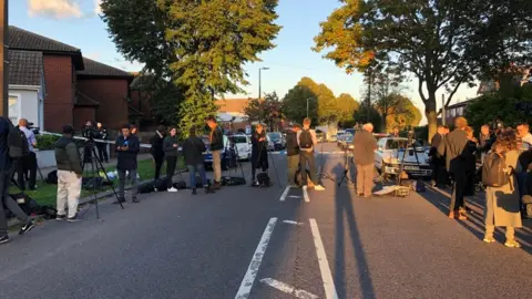 Reporters and people laying flowers have gathered in the normally quiet, residential street