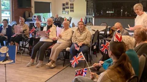 A group of older people, some wearing paper hats and holding Union Jack flags, are sat in a canteen. They appear to be taking part in a group activity such as singing.