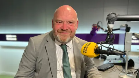 Bill Revans in a suit sat in a radio studio looking at the camera