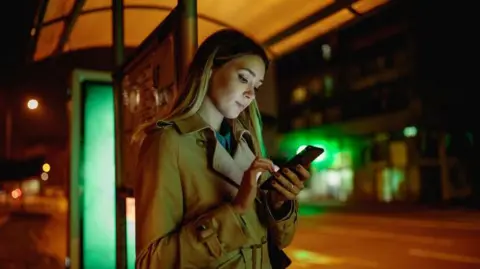 Blonde woman in trench coat at bus stop at night, looking at a phone. 