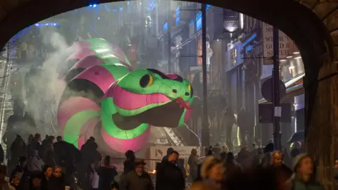 DCSDC Shows a large illuminated snake in green, pink and black going down Shipquay Street, with people in the foreground in front of an arch.