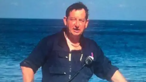 Manche Iles Express Bernard Jambon is looking into the camera. He is wearing blue work clothes and is stood in front of the sea. He has dark hair. He is squinting his eyes as the sun is shining on his face. 