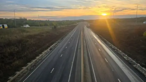 A picture of the A421 carriageway. The photo is taken from a bridge over the carriageway. The road is clear and traffic can be seen heading onto the road via a slip road. The sun can be seen rising in the distance. 