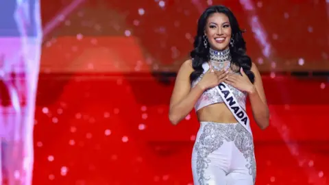 A woman wearing a top and trousers, as well as a sash reading "Canada", holds her hands on her heart