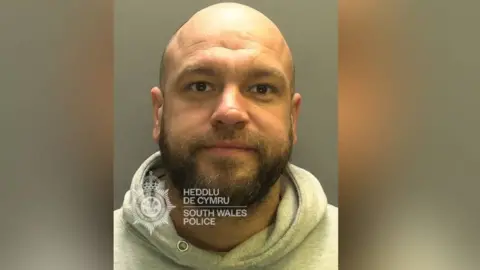 South Wales Police A police mugshot picture of a man with no hair and a beard, wearing a grey hoody 