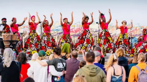 James Bass Photography The African Choir of Norfolk, with its choir members dressed in magenta tops and hairbands, and colourful floral dresses. They are performing on the beach in Great Yarmouth with a vast collection of silk flags made by children fluttering behind them, and a crowd of people watching on. A drummer is to the left of the choir.