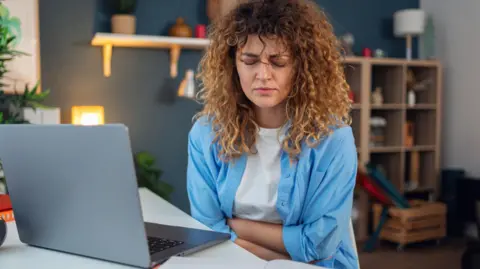 Getty Images Woman with curly brown hair sits at her laptop clutching her stomach in pain. She has closed her eyes and is grimacing.