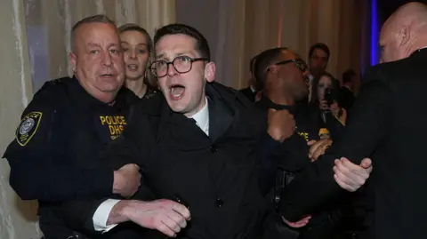 Isaac Burke, who has short, dark hair and is wearing glasses and a black coat, is escorted from the National Building Museum by police and security staff.  Two uniformed officers have their arms under his armpits as he shouts in protest. 