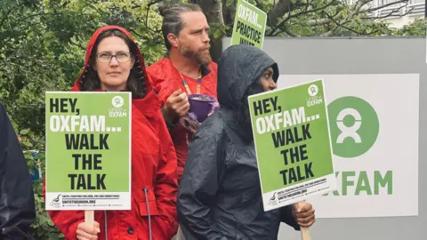 Three protesters with placards next to the Oxfam sign at the charity's headquarters. They are wearing rain coats.