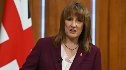 Getty Images Chancellor Rachel Reeves stands in front of a union jack wearing a plum blazer and white V-neck top.