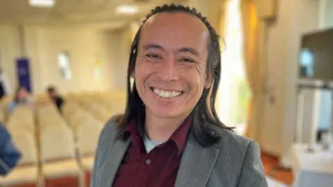 Deputy David Goy is a South Asian man smiling at the camera. He has black shoulder length hair. He is wearing a grey suit and a purple shirt. 