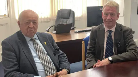 An indoor office environment featuring a wooden meeting table and a workspace behind it. Two men are seated at the meeting table. Both are wearing formal business attire: One is dressed in a textured blazer with a patterned tie and a light-coloured shirt. A small yellow pin is attached to the lapel. The other is wearing a suit jacket, a light shirt, and a patterned necktie. Their hands are resting on the table in front of them.