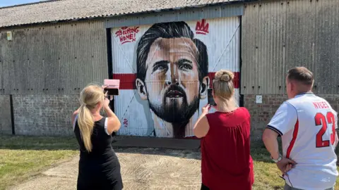 John Devine/BBC Jade and Rachael taking pictures of the mural of Harry Kane, with Nathan Murdoch looking on. He is wearing a replica white England shirt with the number 23 and "NYCES" on it.