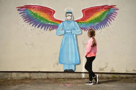 Oli Scarff / Getty Images A girl walks past a mural of a doctor with rainbow angel wings