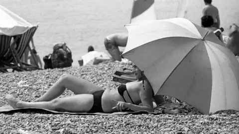 A woman lying on a towel on a pebble beach wearing a bikini in a black and white photograph. She is shaded by a large umbrella. There are several other people set up on the beach in the background.