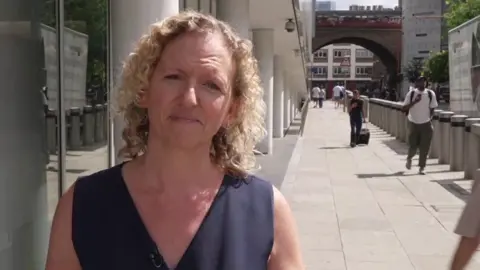 Lucy Hayward-Speight stands on a pedestrian walkway between modern buildings. Lucy Hayward-Speight is wearing a dark sleeveless top. Railway arches, office buildings and several pedestrians can be seen in the background.