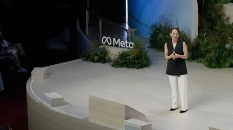 Gleit wears a sleeveless black blazer, white trousers and black shoes as she addresses the audience on-stage at an event in London. A sign bearing Meta's logo is surrounded by foliage behind her on the stage.