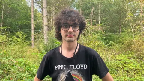 Teenager with black curly hair standing in a wood wearing a Pink Floyd t-shirt.