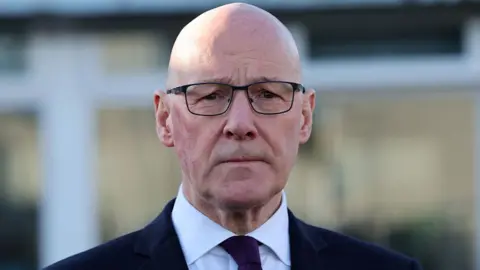 John Swinney, a bald man wearing glasses, a dark suit, purple tie and a white shirt, stares into the camera with a neutral expression