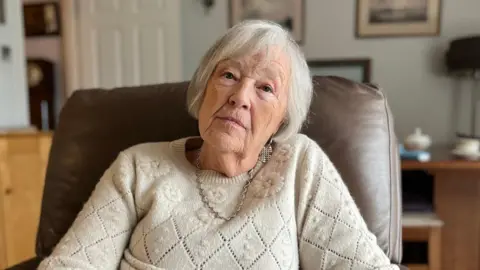 A grey-haired woman sits in a leather arm chair and looks solemn into the camera. She has a beige jumper on.