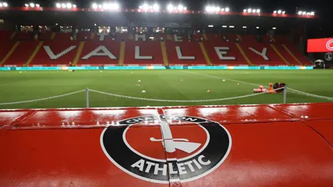 A view of Charlton Athletic's empty football stadium, The Valley, at night under bright floodlights. In the foreground, the top of a red dug-out seat or tunnel structure is prominently featured, displaying the circular Charlton Athletic crest with a hand holding a sword. Beyond the green pitch, red stadium seating spells out "The Valley" in large white lettering across the main stand.