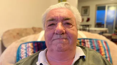 A man with white hair and dark bushy eyebrows sits inside a living room, giving a closed-mouth smile to the camera.