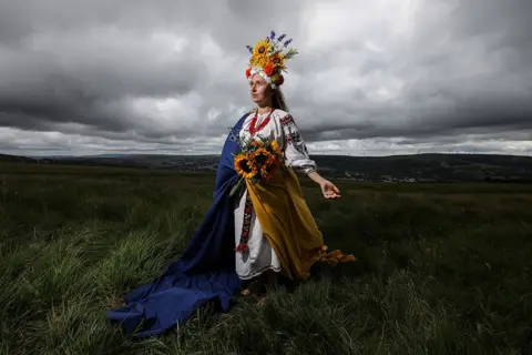 Joann Randles Stood barefoot, in a traditional Ukrainian embroidered dress and Vinok, proudly draped in the Ukrainian flag, Olha Boyko, celebrates Ukraine Independence Day