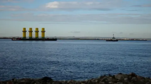 Getty Images Four monopile structures used as the foundation of wind turbines are transported on a barge towed by a tug boat out of the mouth of the River Tees in Redcar in 2020