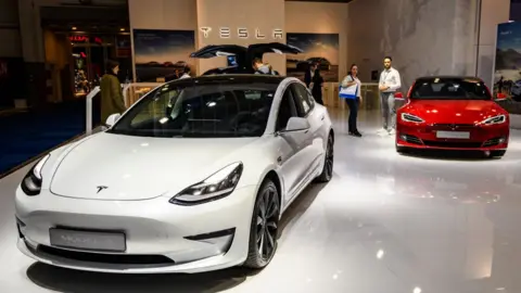 Getty Images A white Tesla model 3 is seen on the left and a Red Model S on the right on a show floor