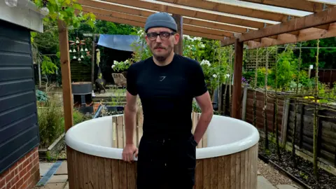 A man in an all black running outfit and beanie hat standing outside in a garden.