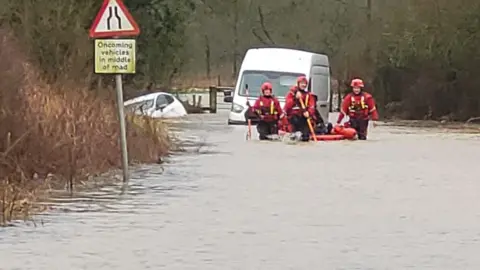 Three rescue workers can be seen wading through water at knee height - a submerged van and car can be seen in the background