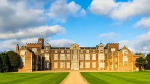 Burton Constable A large Elizabethan red-brick mansion, built in a H-shape with three stories, cupolas and, to the left, a tower with battlements. It sands in green grounds with a gravel driveway, under a blue sky.