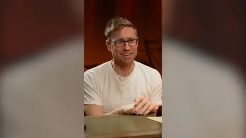 Russell Howard sits behind a desk with his hands on the table. He has short blond hair and wears a white T-shirt