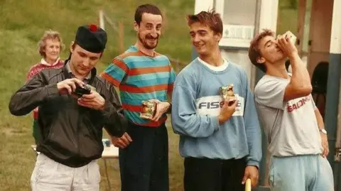 Molly Gill Four men in baggy sports clothes share a joke as they drink from cartons of juice. They are standing at the foot of a grass hill.