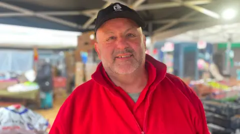 A man in a red top and mainly black cap is looking at the camera. The market setting behind him is blurred.