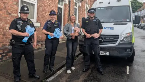 Three uniformed police officers - a man and a woman - and another woman wearing jeans and a grey fleece sanding in front of a marked police van parked on a terraced street. The two on the left are holding blue hand-shaped leaflets with printing on them and the other two leaflets depicting hairy hands.