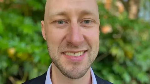 Andrew Sinclair/BBC Ed Birkett is smiling at the camera. He is wearing a navy blue suit jacket over a light blue shirt.